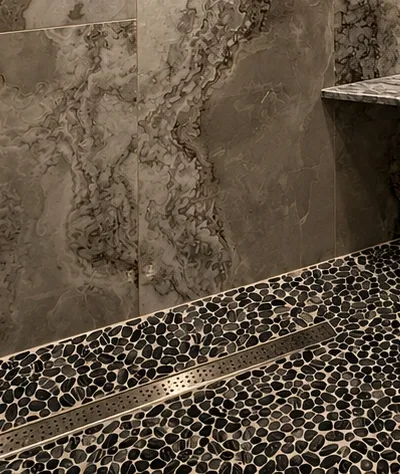 Detail of a pebble mosaic shower floor pitched to a linear drain, meeting the veined wall tile at a clean junction.