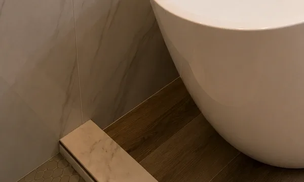 Detail where a tiled curb and mosaic edge meet a plank floor beside a freestanding tub.